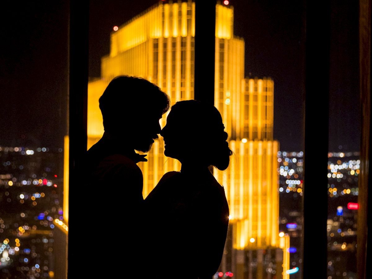 A silhouetted couple stands in front of a brightly lit cityscape at night, framed by a large window in this image.