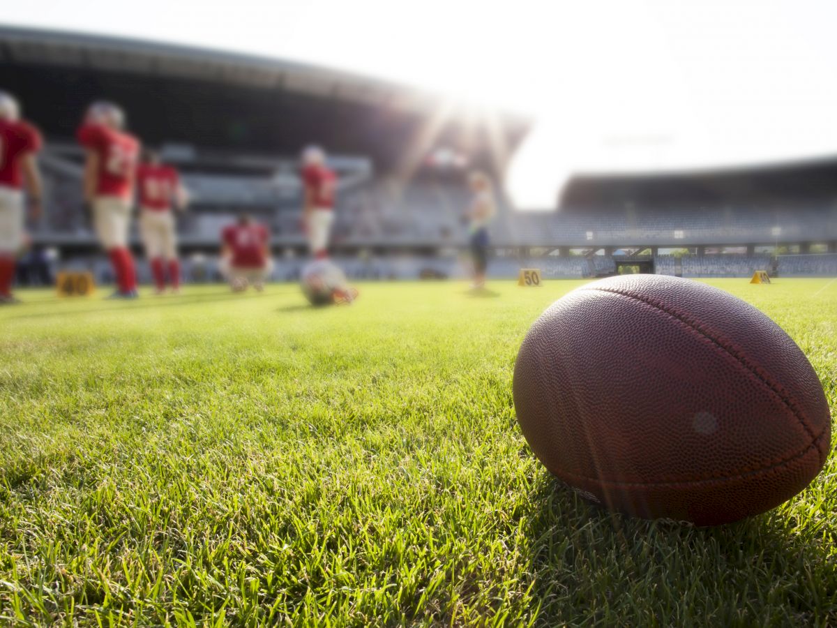 A football lies on the grass near a group of players wearing red uniforms, with a stadium and sunlight in the background.