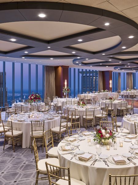 A beautifully decorated banquet hall with round tables set for an event. Large windows offer a city view, and elegant lighting adorns the ceiling.