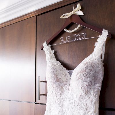 A white lace wedding dress hangs on a wooden hanger with a wire showing the date "3.13.2021" above a pearl necklace, against a wooden cabinet.