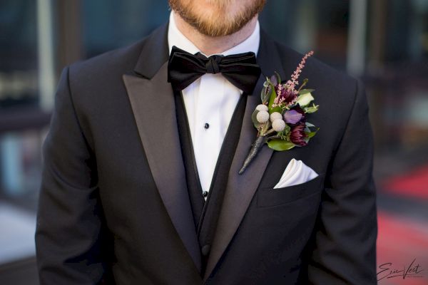 A person wearing a black tuxedo with a white dress shirt and black bow tie, accessorized with a boutonniere featuring flowers and greenery.