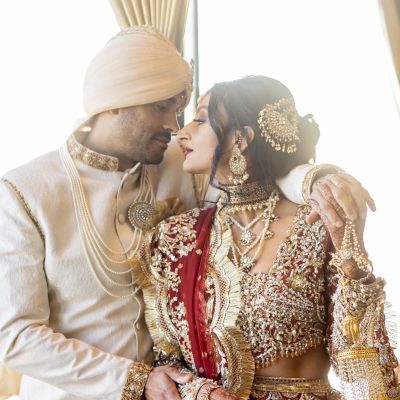 A couple dressed in traditional Indian wedding attire shares an intimate moment, with the groom in white and the bride in a red and gold outfit.