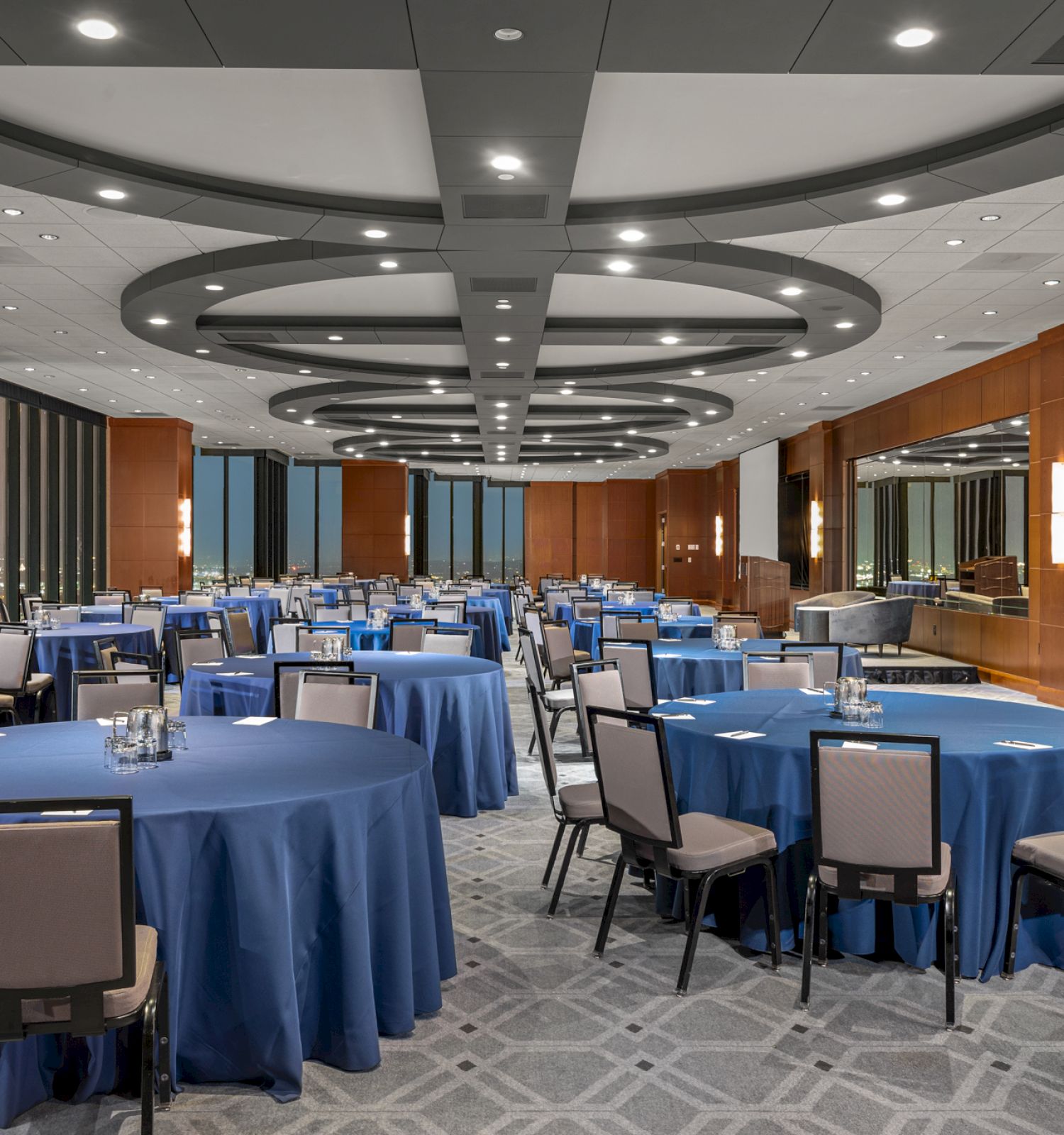 A conference room with round tables covered in blue cloth and surrounded by chairs, featuring a modern ceiling and wall decor.