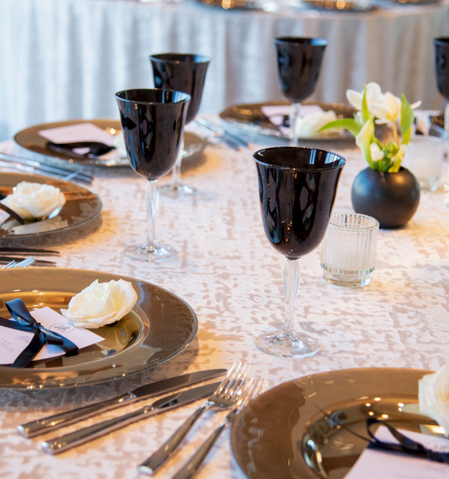 A table set elegantly with black goblets, gold-rimmed plates, white flowers, and neatly arranged cutlery, creating a formal dining ambiance.