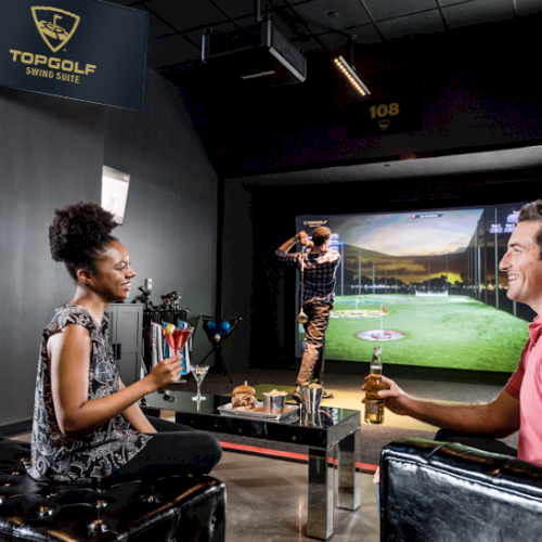 People enjoying a virtual golfing experience at Topgolf Swing Suite, with a man swinging a golf club and others relaxing with drinks.