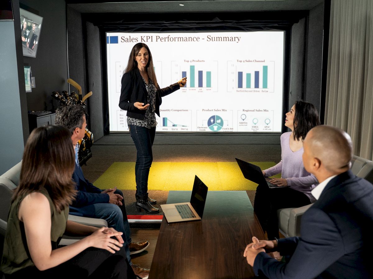 A woman is presenting a "Sales KPI Performance - Summary" on a screen to four colleagues seated around a table with laptops open.