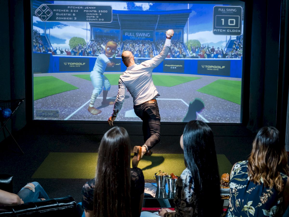 A man is playing a baseball video game on a large screen, while three people watch from behind in a dark indoor setting.