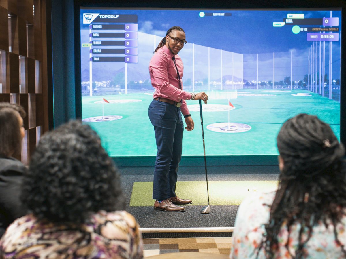 A person holding a golf club stands in front of a virtual golf screen while others watch. The setting appears to be an indoor golf simulator.