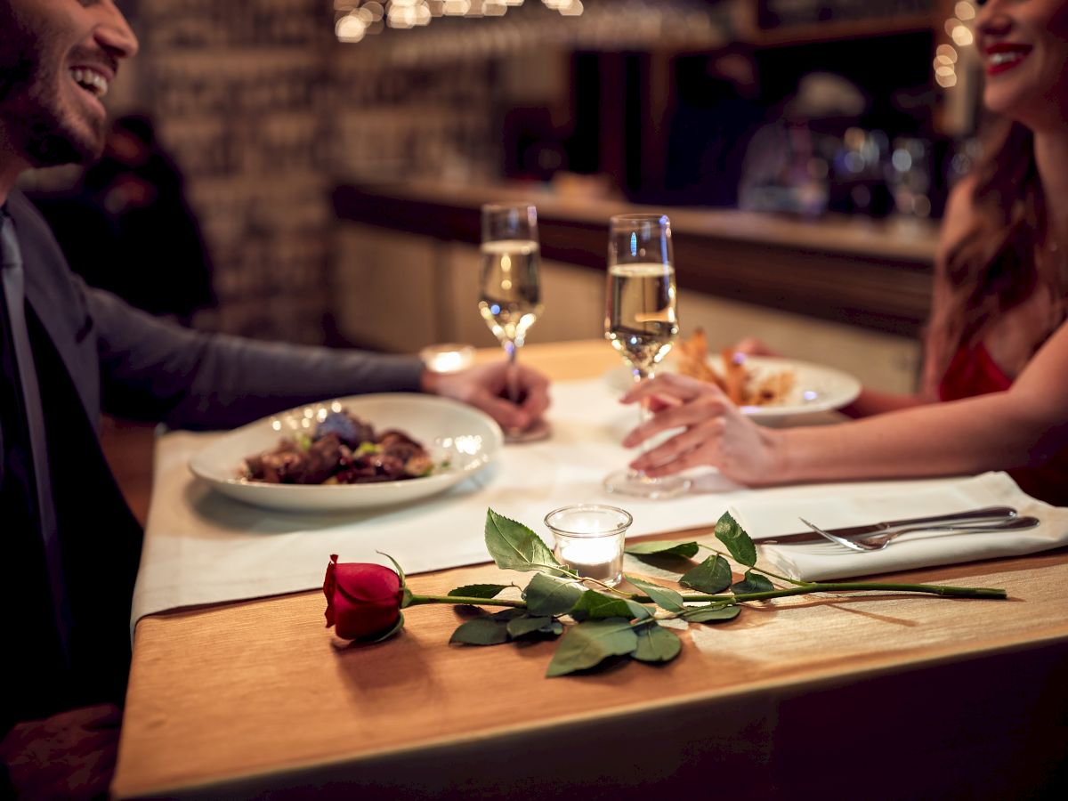 A couple is dining at a restaurant, with wine glasses, plates, and a rose on the table, creating a romantic atmosphere.