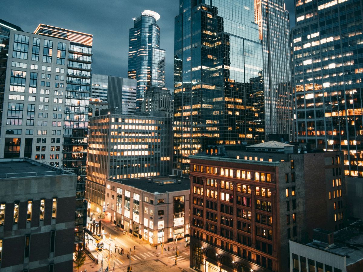 A cityscape at dusk showing tall buildings with lights on, under a cloudy sky, with streets illuminated below, creating a bustling urban atmosphere.