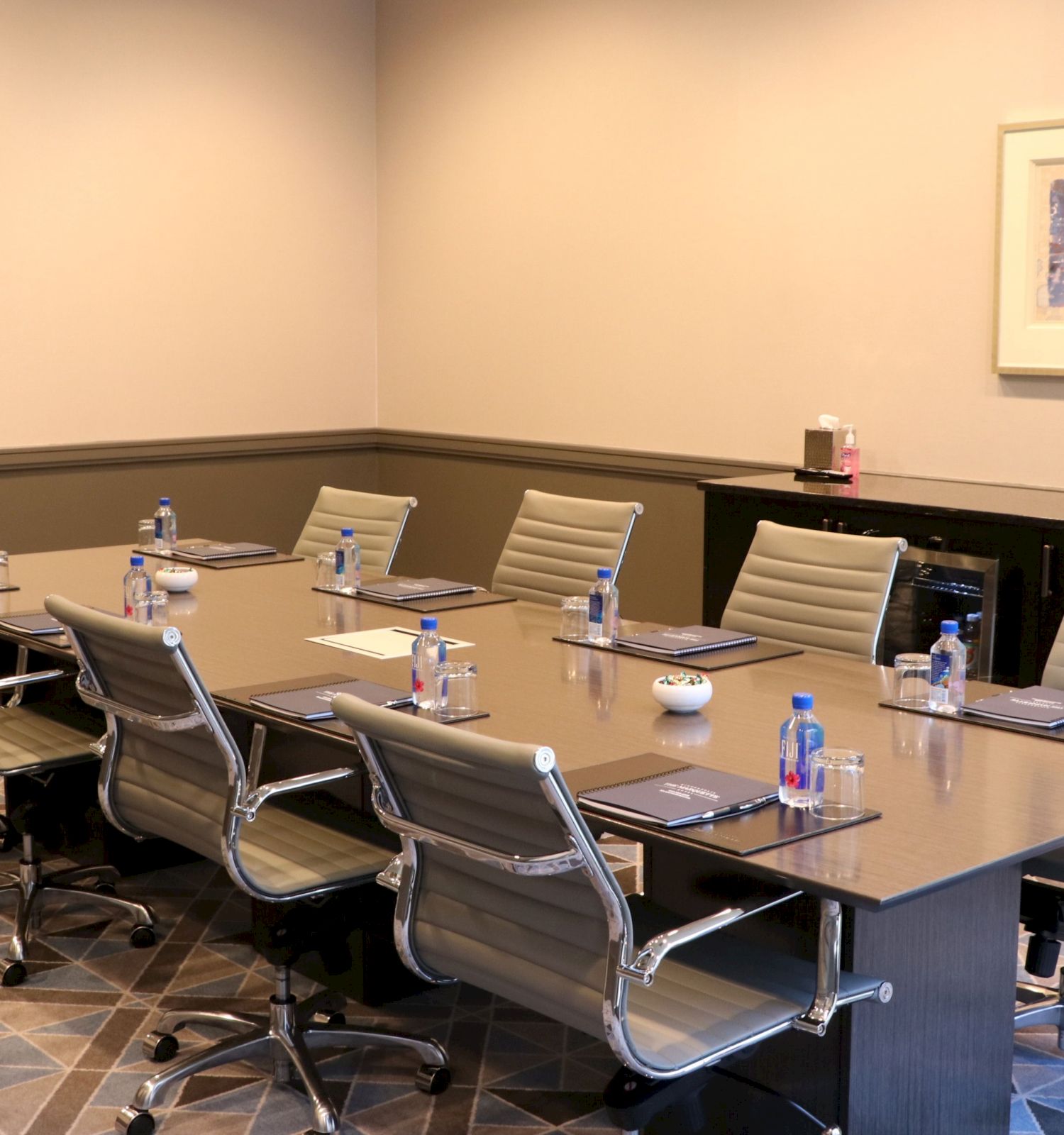 A conference room with an oval table, office chairs, water bottles, notepads, a wall-mounted TV, and a sideboard with refreshments and paper supplies.