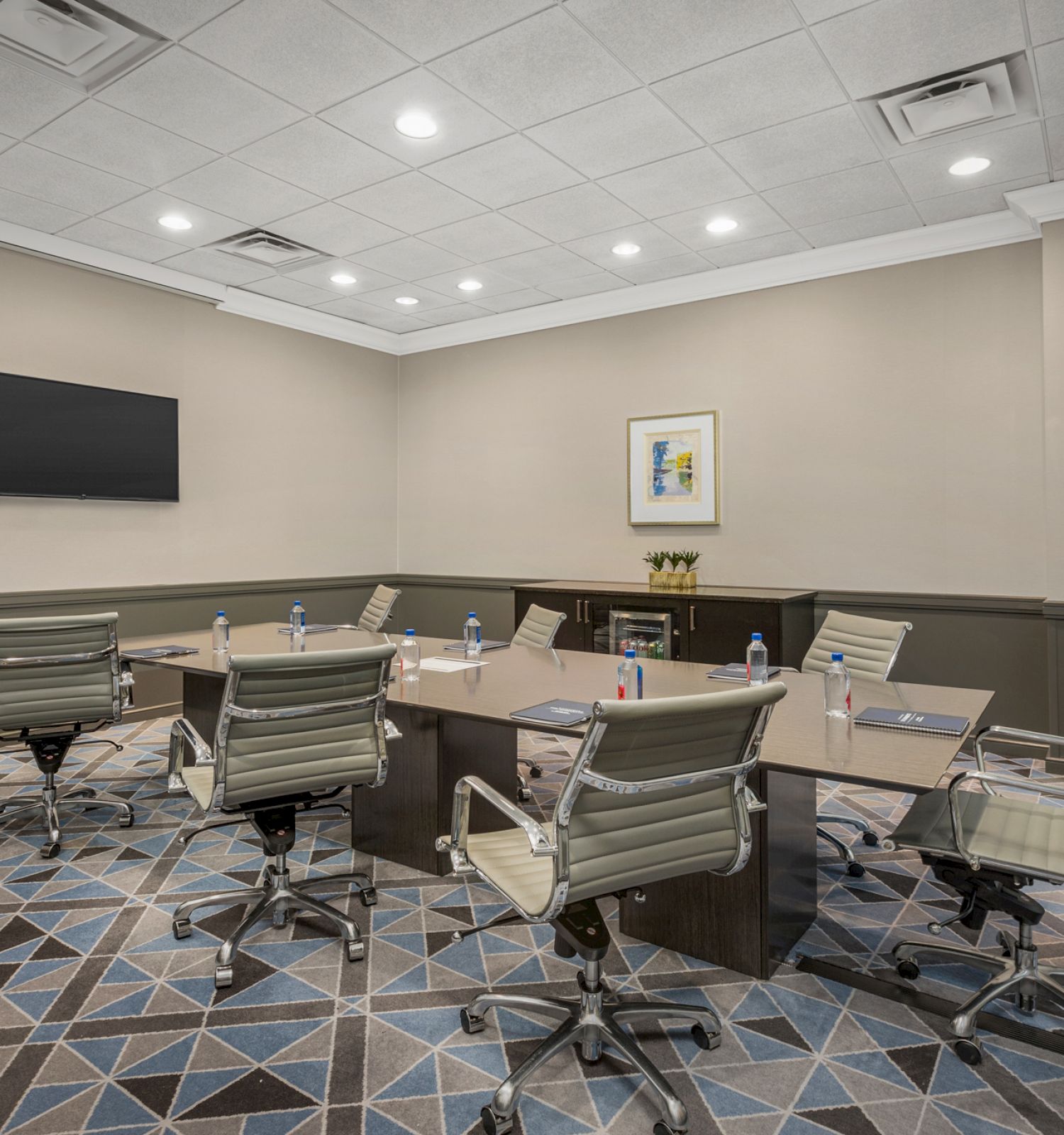 A modern conference room with a rectangular table, six swivel chairs, water bottles, notepads, a TV on the wall, and a piece of art above a cabinet.