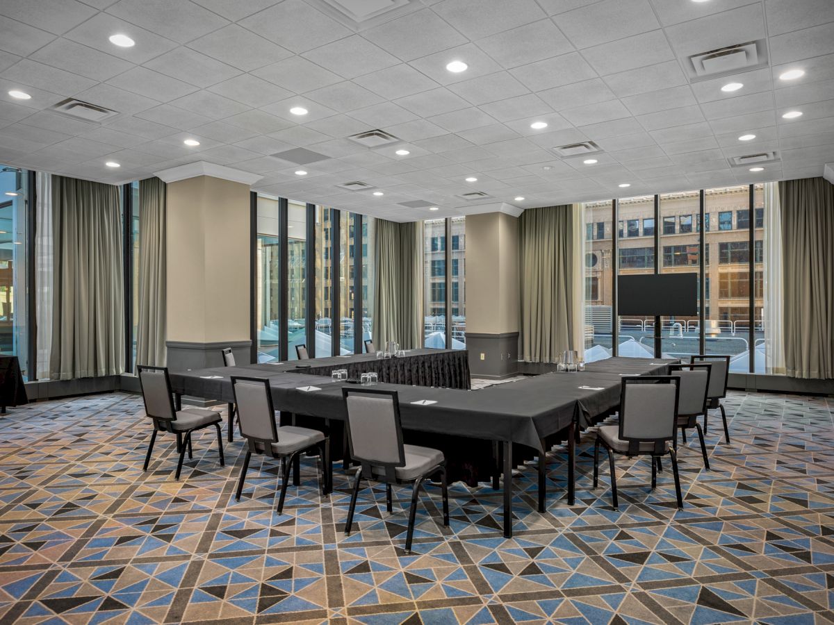 A modern conference room with large windows, curtains, a U-shaped table setup with chairs, and a screen at the far end.