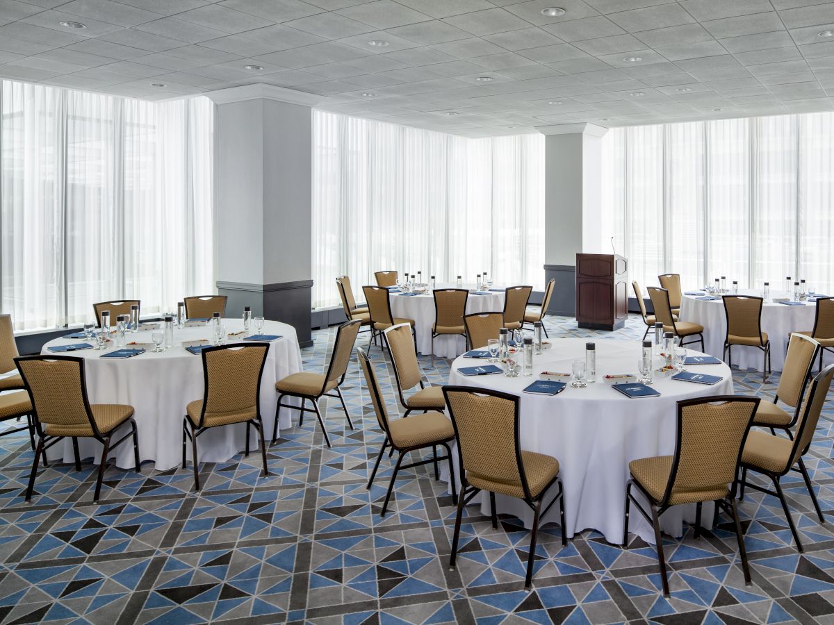 The image shows a conference room set up with round tables and chairs, each table having writing materials and water bottles.