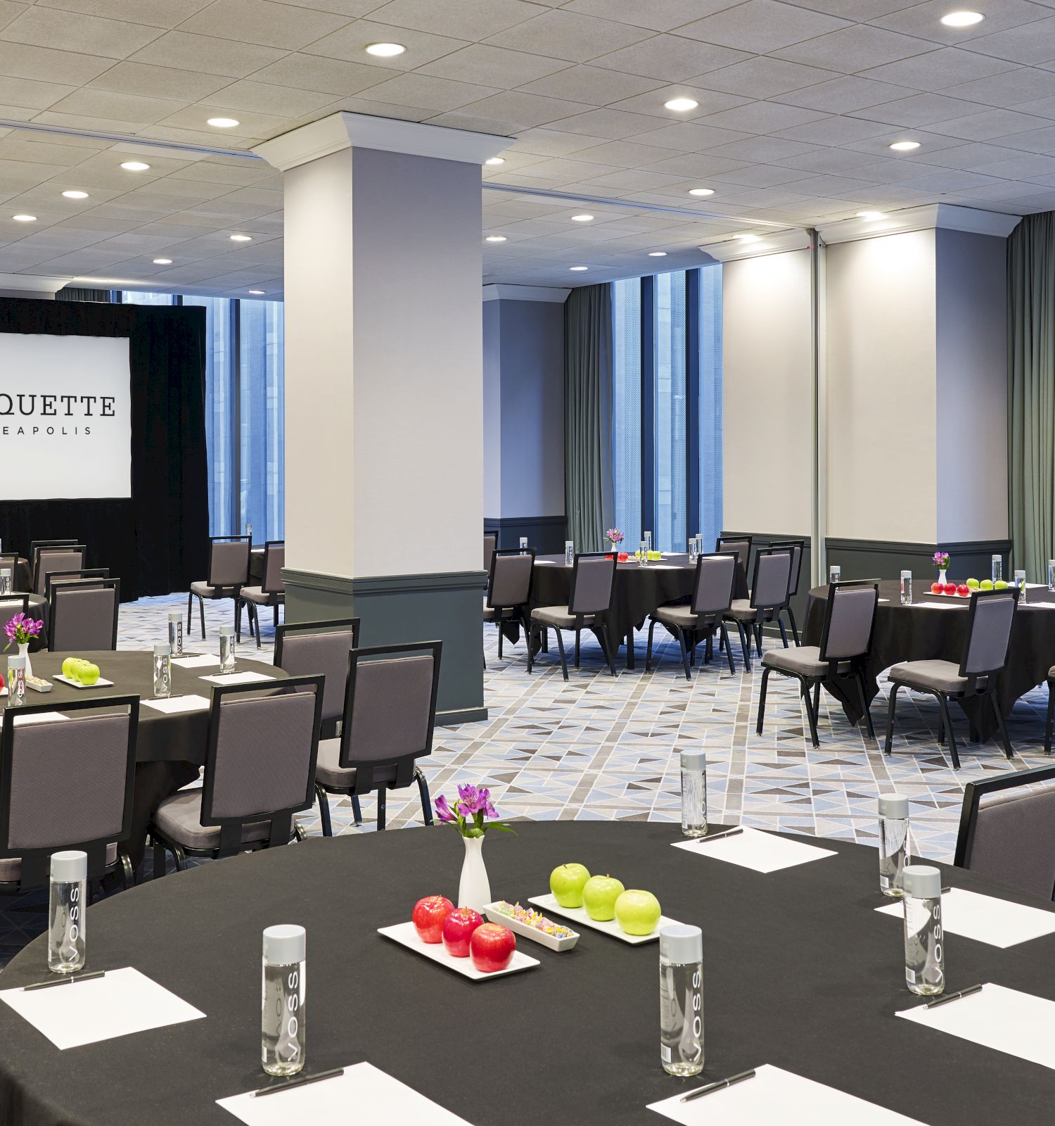 A conference room with round tables, chairs, water bottles, notepads, and colorful centerpieces. A screen in the back displays 