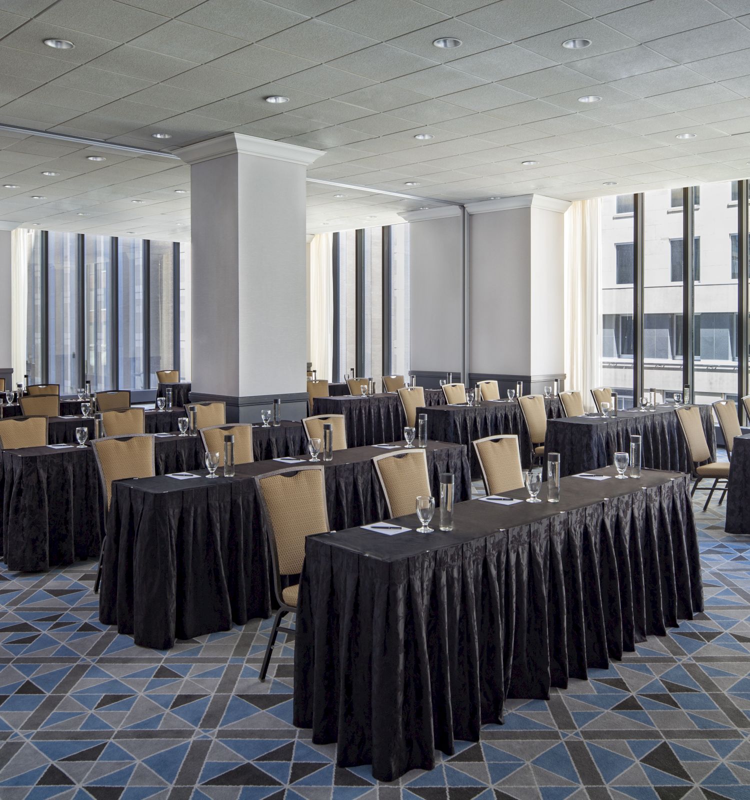 A conference room with multiple tables and chairs arranged in rows, with glasses, notepads, and water bottles on the tables, and large windows in the background.