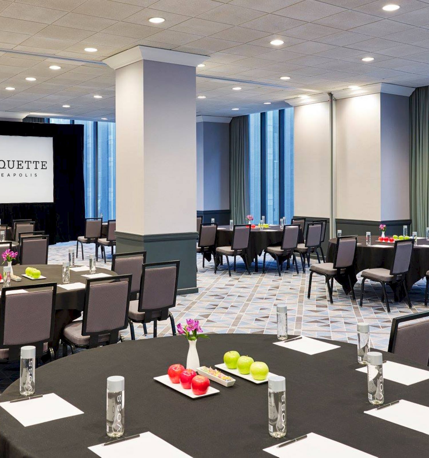 The image shows a conference room setup with round tables and chairs, a projector screen displaying "Marquette," and tables with notepads, water bottles, and apples.