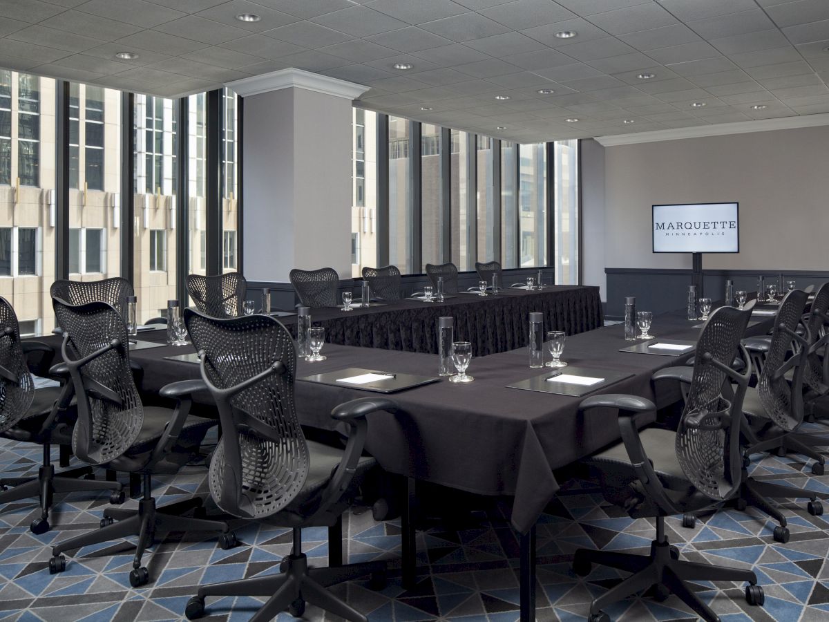 A modern conference room with long tables, black chairs, notepads, glass cups, and a TV screen displaying the 