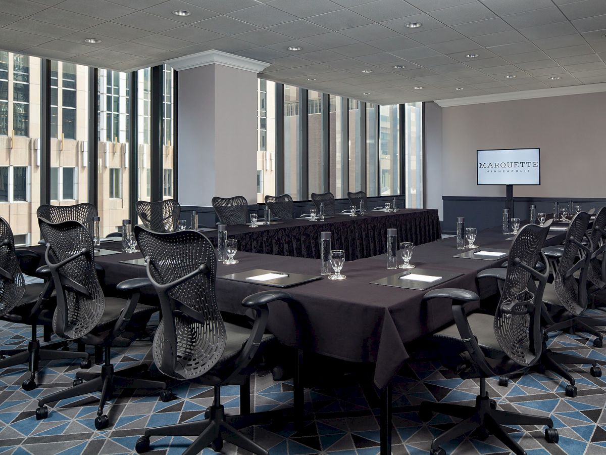 A modern conference room with black chairs, tables arranged in a U-shape, glasses, paper, and pens, and a TV displaying 