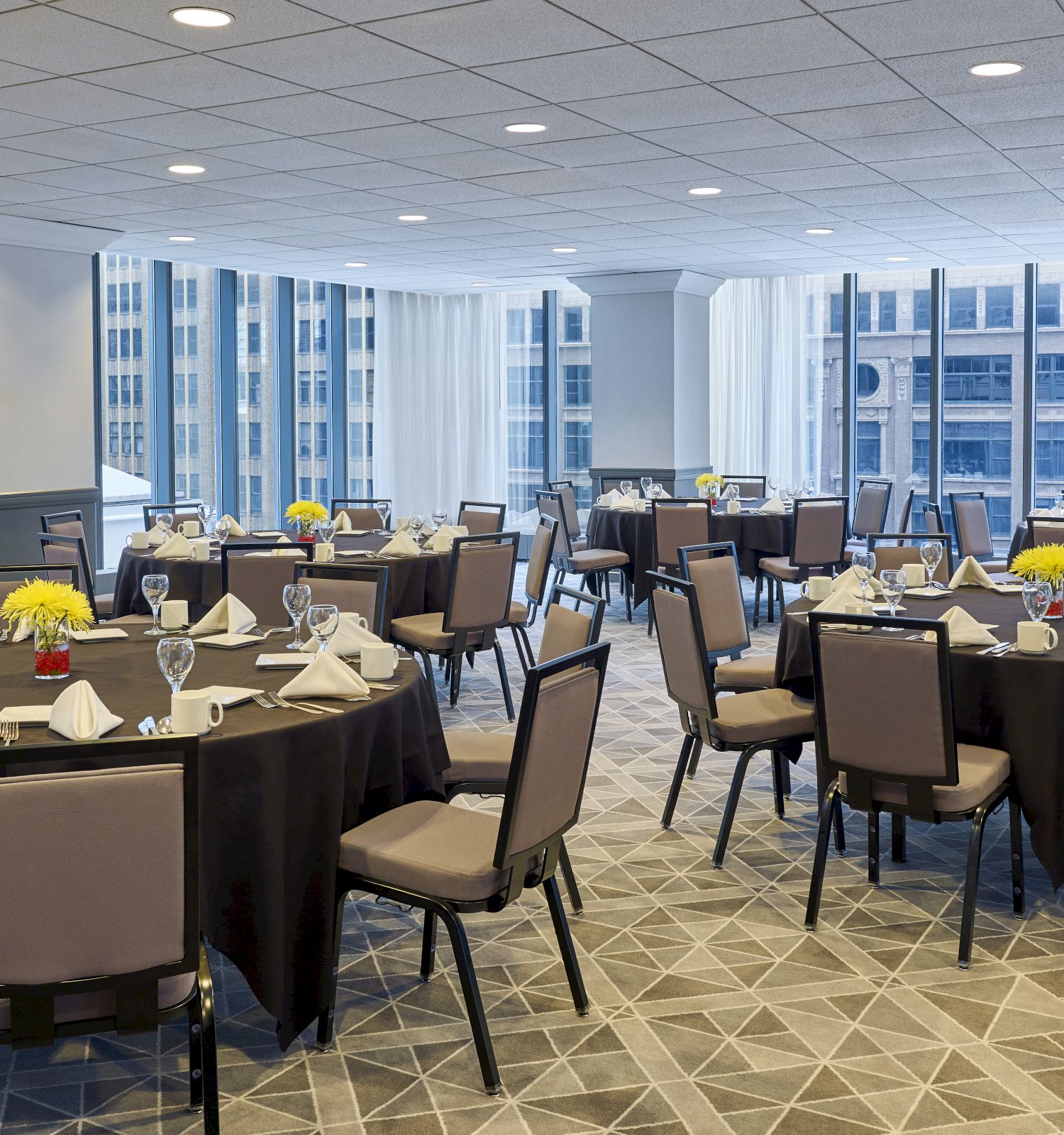 A spacious, elegantly set dining area with round tables, chairs, neatly folded napkins, and floral centerpieces against a backdrop of large windows.