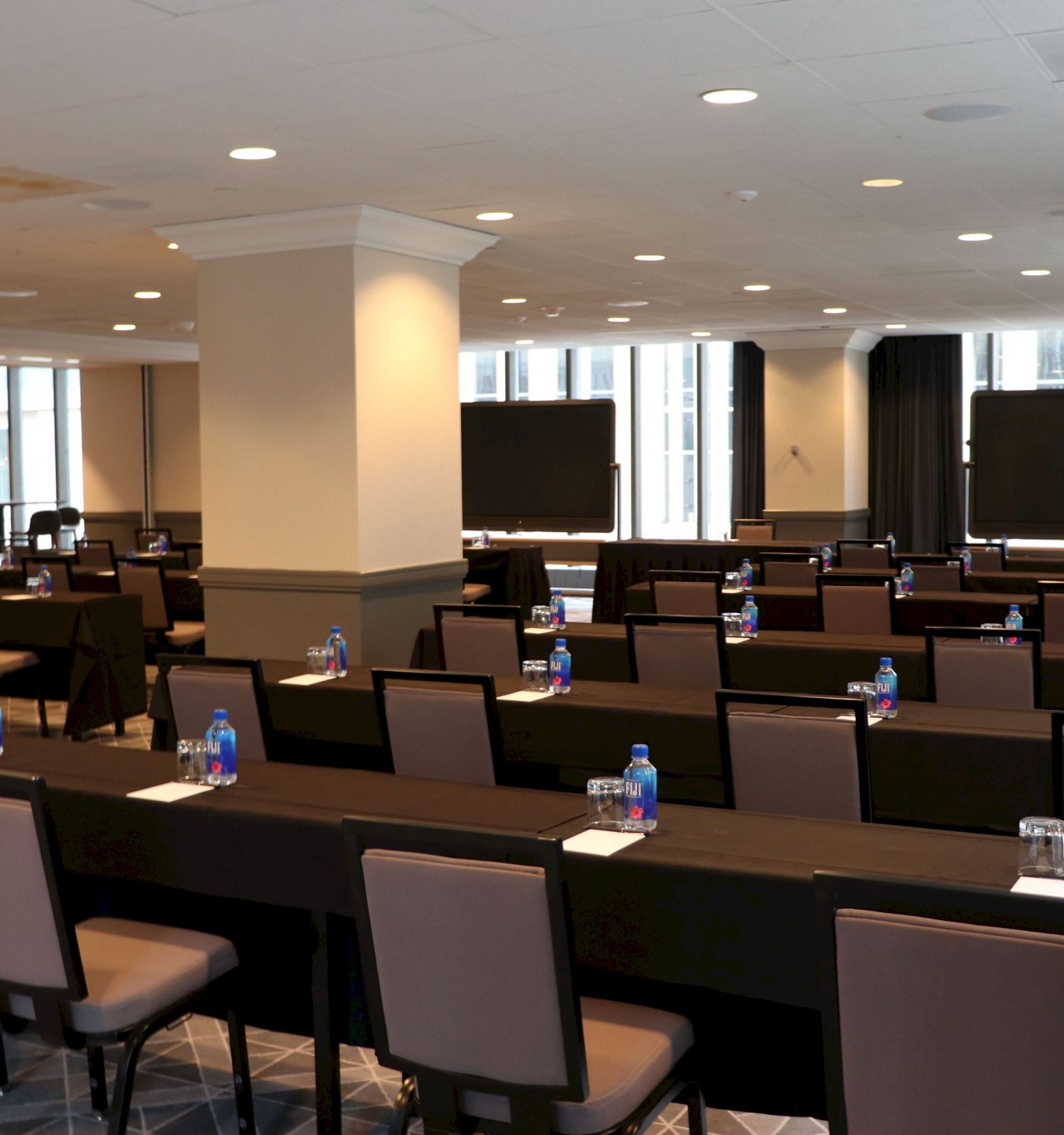 A conference room setup with rectangular tables and chairs arranged in rows facing two large screens. Bottles of water and notepads are on the tables.