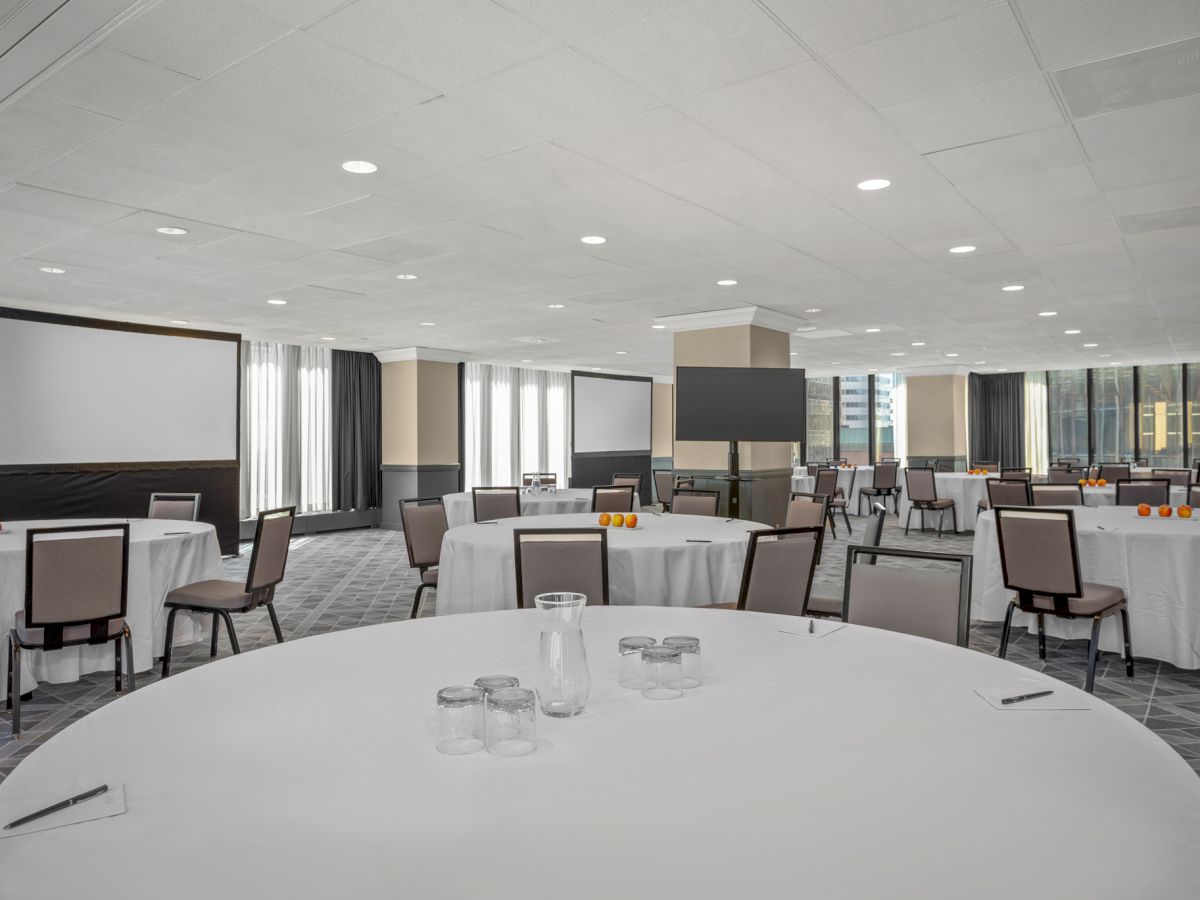 The image shows a spacious conference room with round tables and chairs, two large screens, and natural light coming in through large windows.