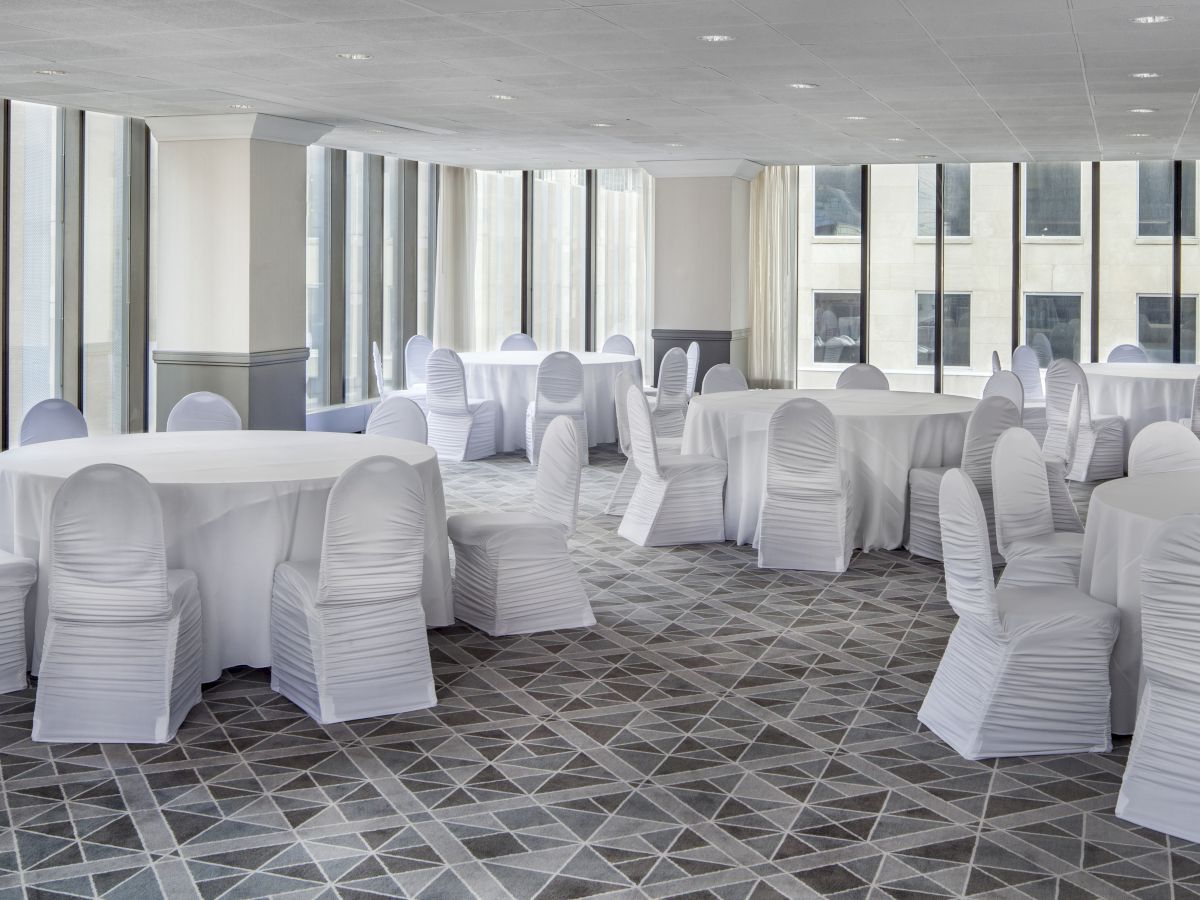 The image shows a banquet hall with several round tables and chairs covered in white fabric, set up near large windows.