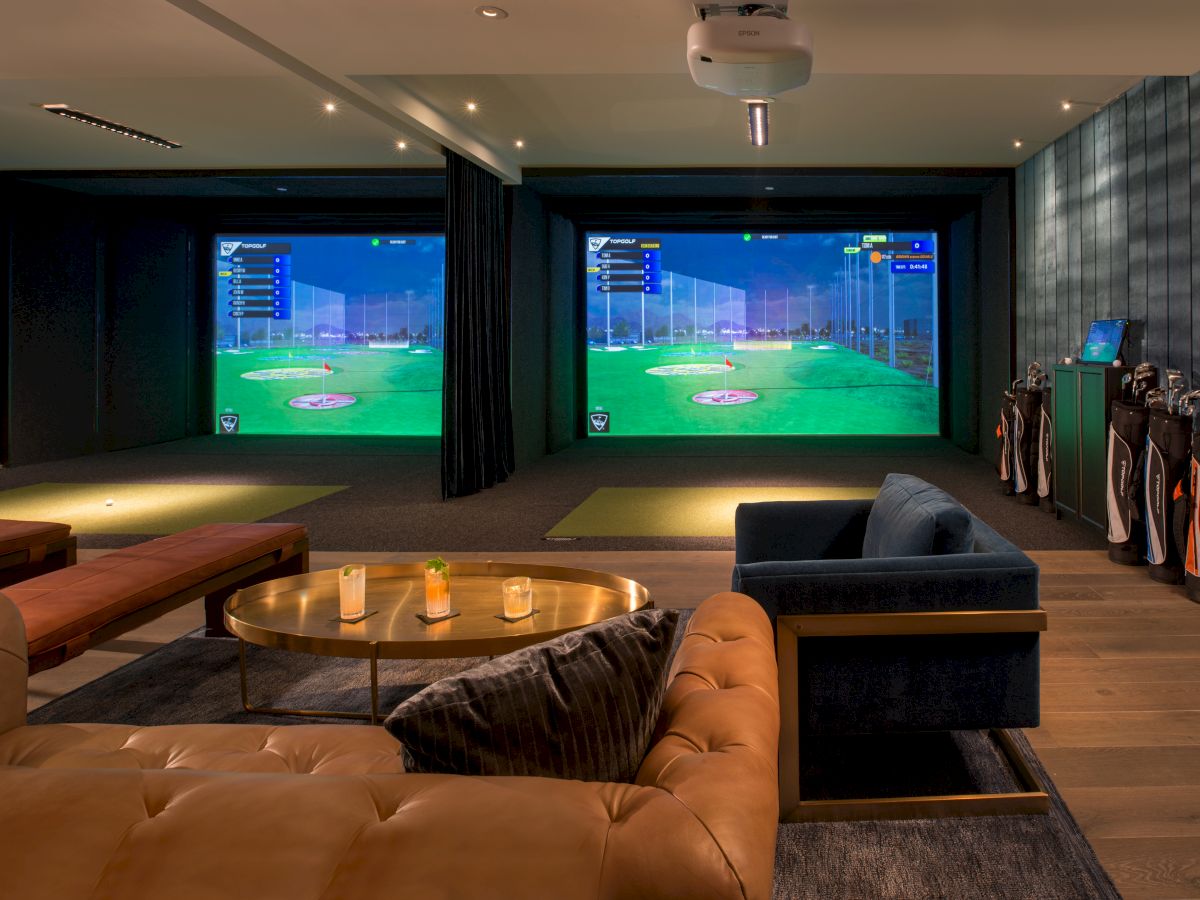 The image shows an indoor golf simulator room with sofas, candles, a round table, and multiple sets of golf clubs on the right.