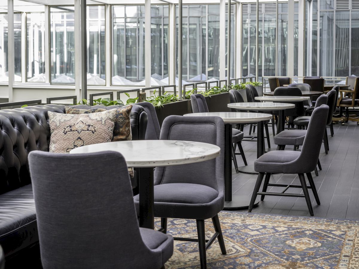 A modern seating area featuring marble tables, gray upholstered chairs, and a black tufted sofa with throw pillows, set in an airy, windowed space.