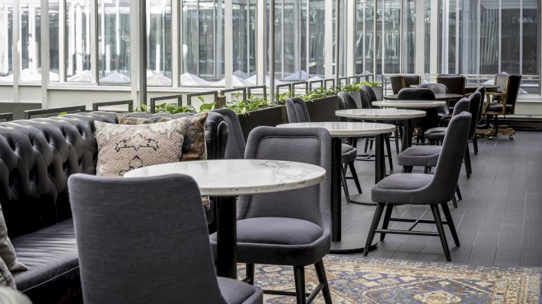 A modern seating area featuring marble tables, gray upholstered chairs, and a black tufted sofa with throw pillows, set in an airy, windowed space.