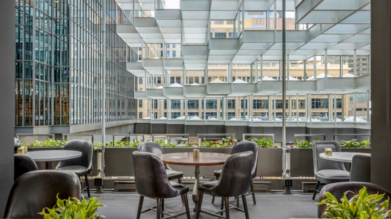 An outdoor terrace with tables and chairs, surrounded by glass buildings and decorated with green plants, is shown in the image.