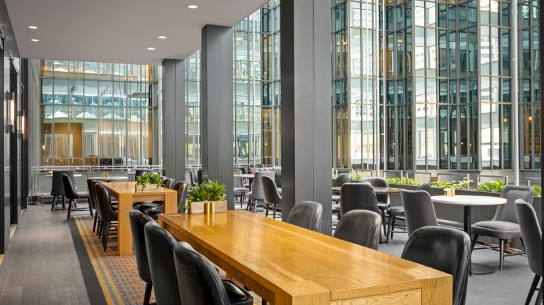 A modern dining or meeting area with wooden tables, black chairs, and glass walls, featuring potted plants and high ceilings in a well-lit space.