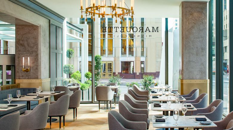 The image shows a modern restaurant with elegant seating, large windows, and a chandelier. The name 