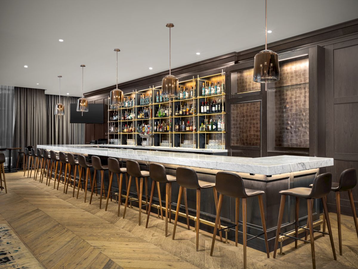 A modern bar with a marble countertop, high stools, and shelves stocked with bottles. It has pendant lighting and wooden flooring.