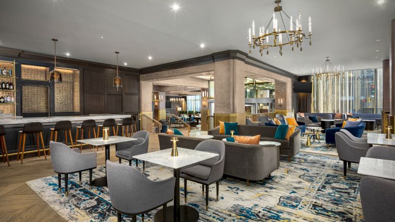 A modern, elegant lounge area with grey and blue furniture, a bar with high stools, and a chandelier, featuring a contemporary design and decor.