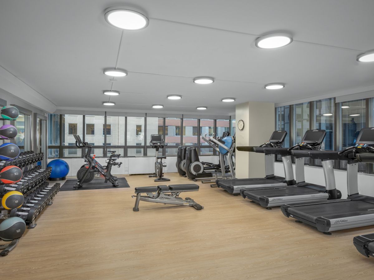 This image shows a modern gym with treadmills, exercise bikes, weights, and other fitness equipment. The room has large windows and bright lighting.