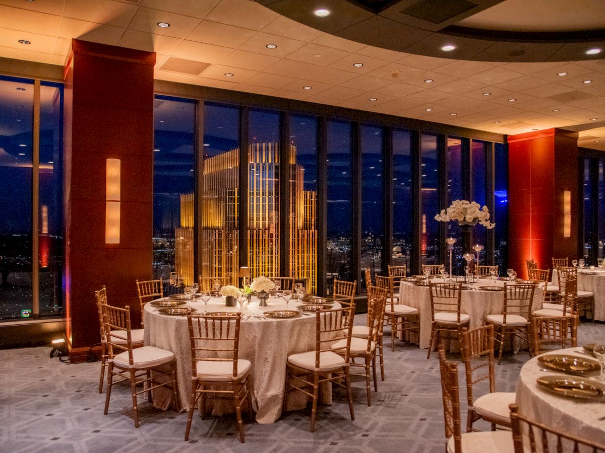 The image shows an elegantly set dining area with round tables and chairs, large windows, and a city view at night.