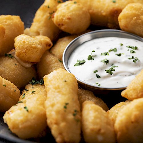 The image shows a plate of fried cheese curds with a small container of dipping sauce, garnished with herbs, set against a dark background.