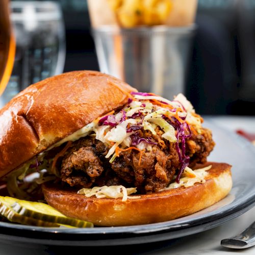 A fried chicken sandwich with coleslaw on a plate with pickles, accompanied by a drink and a small container of sauce on the side.