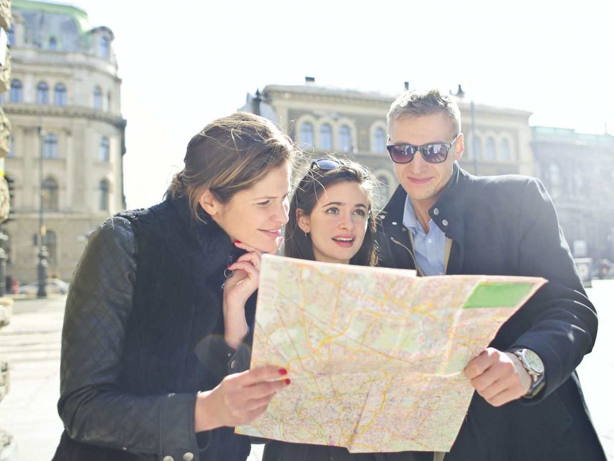 Three people are outdoors, looking at a map together with buildings in the background and sunlight shining through.