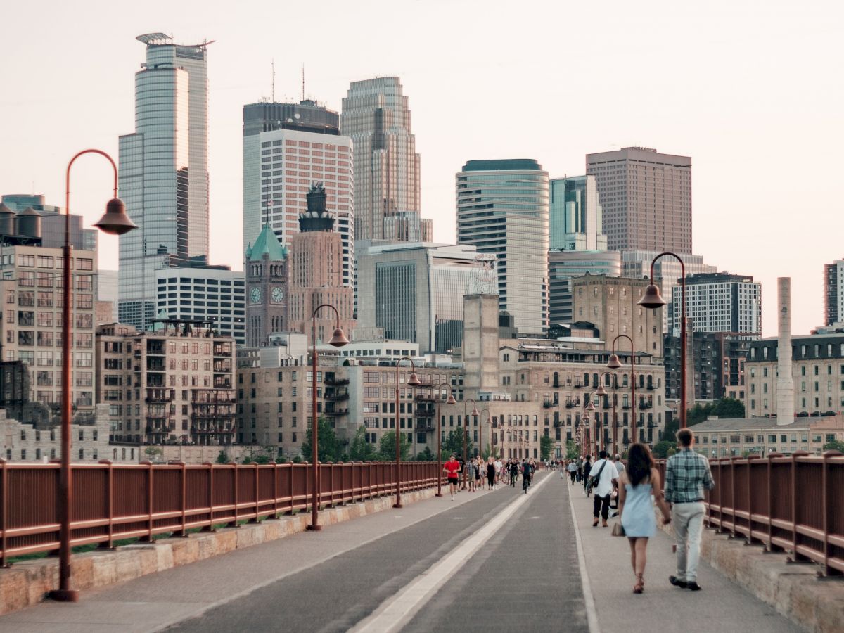 The image shows a cityscape with skyscrapers. People are walking on a bridge with street lamps, leading towards the urban area in the background.