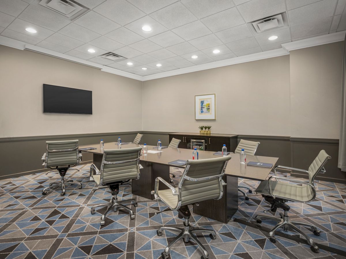 A modern conference room with a rectangular table, six chairs, a wall-mounted TV, and office supplies on the table.