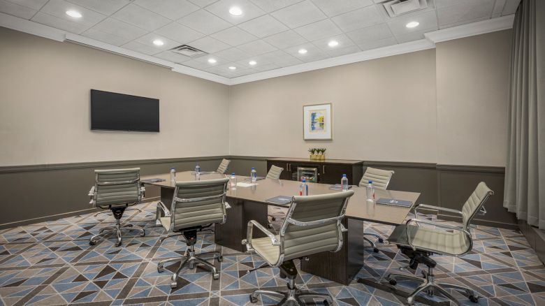 A modern conference room with a rectangular table, six chairs, a wall-mounted TV, and office supplies on the table.