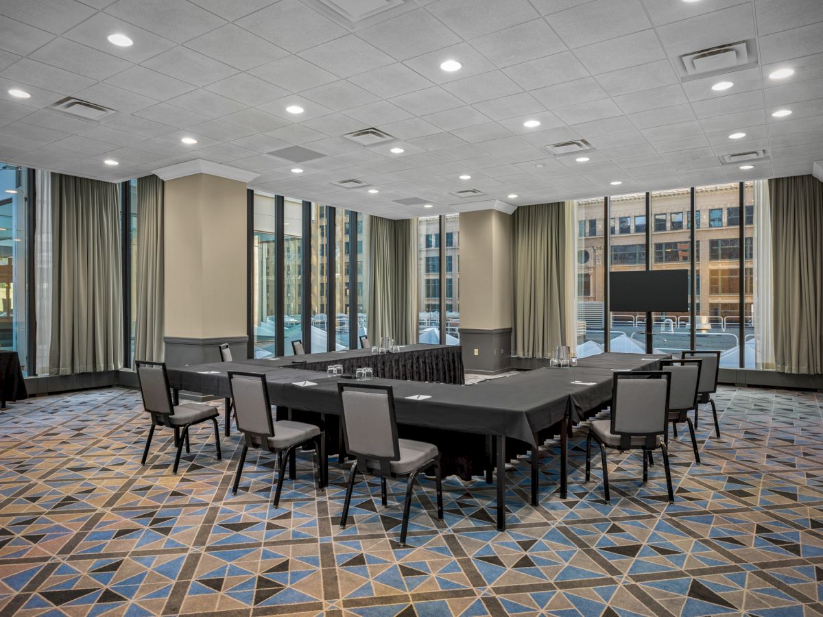 A modern conference room features large windows, arranged with tables and chairs in a U-shape, with a TV screen and patterned carpet flooring.