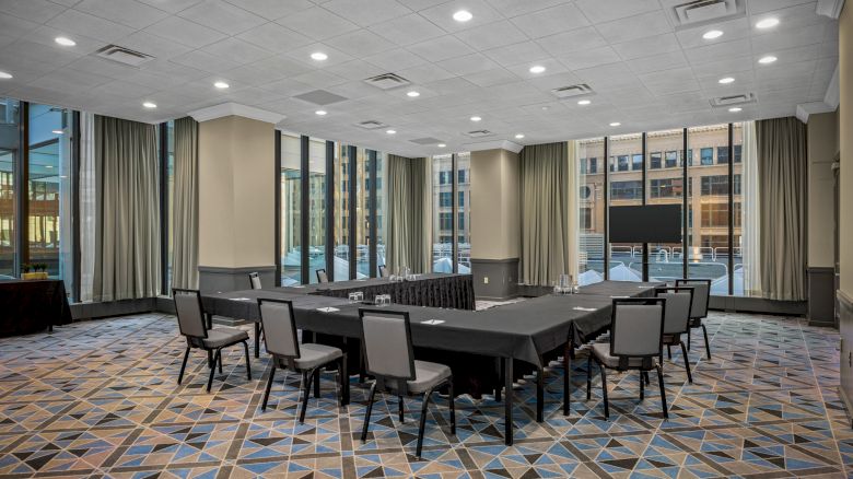 A modern conference room features large windows, arranged with tables and chairs in a U-shape, with a TV screen and patterned carpet flooring.