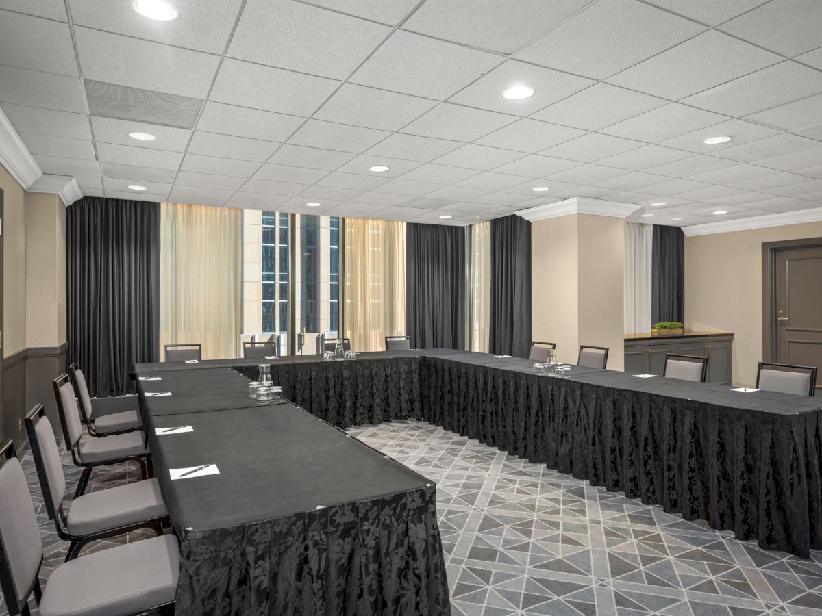 A conference room with a U-shaped table setup covered in black cloth, chairs, notepads, water glasses, and a window in the background.