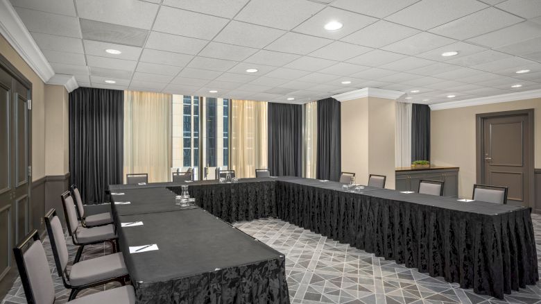 A conference room with a U-shaped table setup covered in black cloth, chairs, notepads, water glasses, and a window in the background.