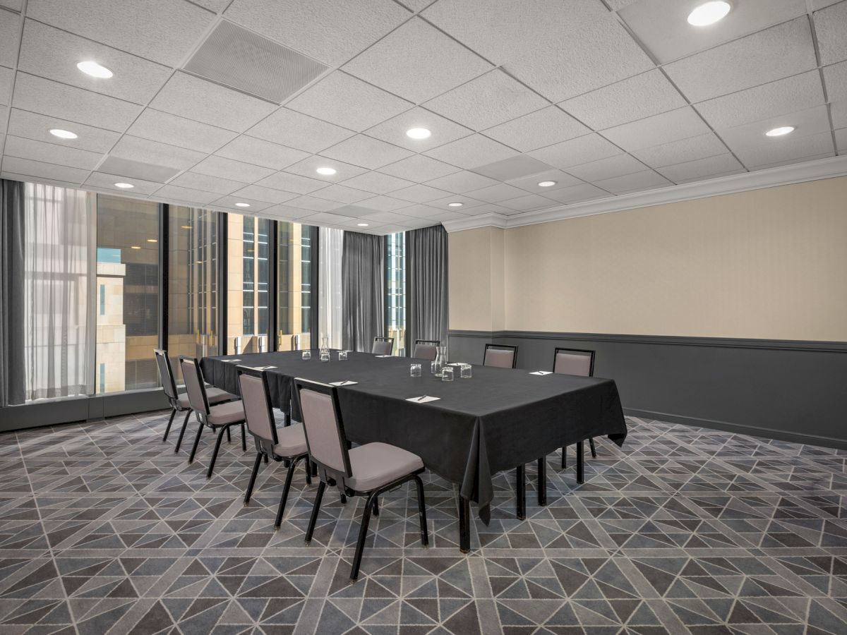 A conference room with a rectangular table covered with a black tablecloth, surrounded by eight chairs, and large windows with city views.