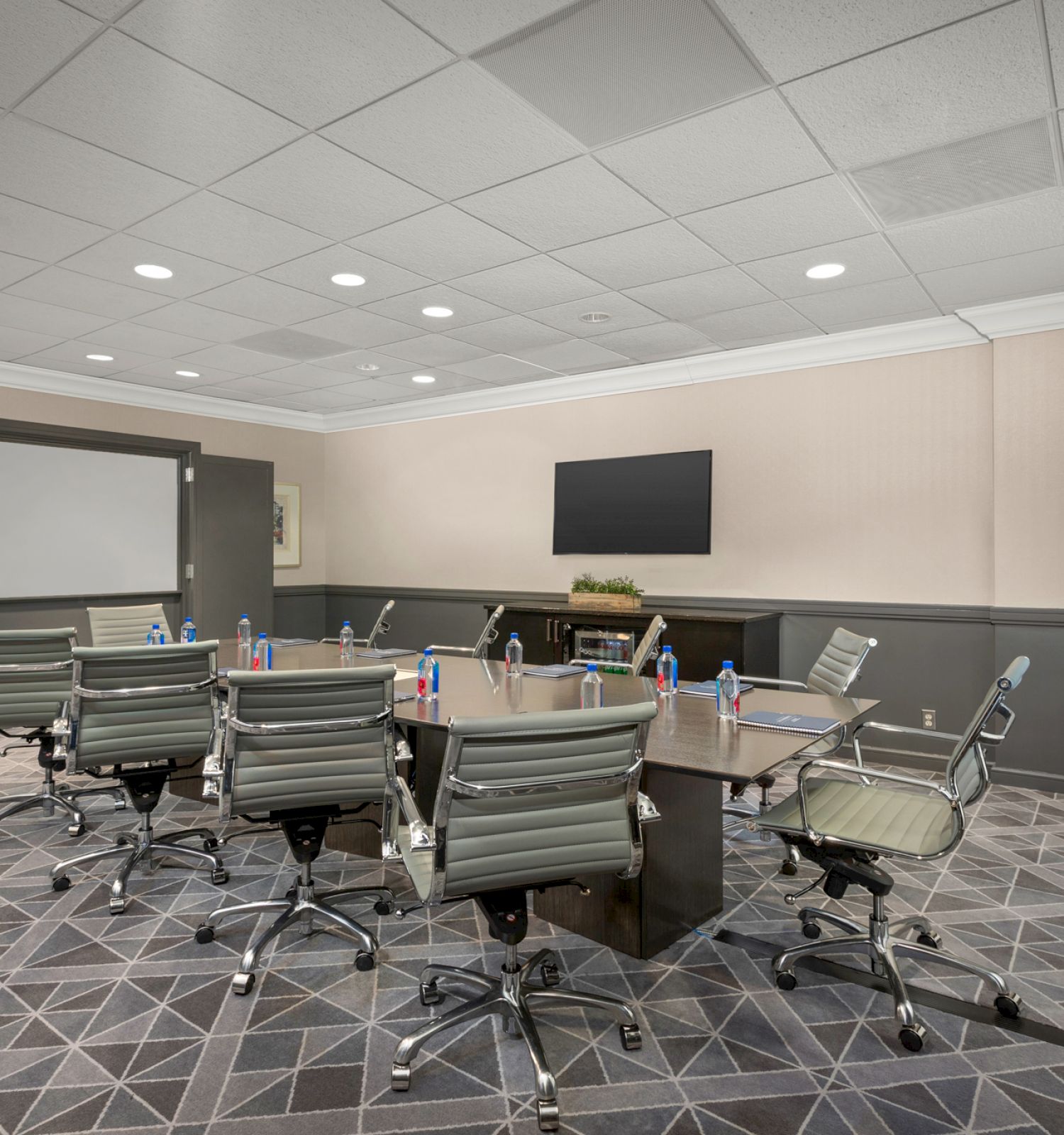 The image depicts a conference room with a rectangular table, surrounding chairs, a TV, and a whiteboard. Bottled water and notepads are on the table.