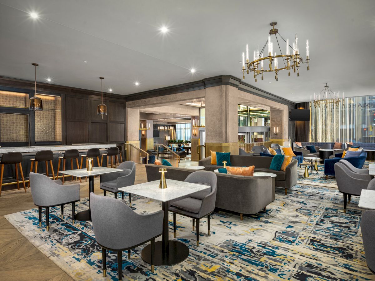 A modern lounge area with comfortable seating, a bar with stools, stylish décor, elegant chandeliers, and a colorful patterned rug on the floor.